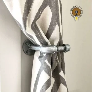 Silver L-shaped industrial pipe curtain holdback holding a gray and white curtain against a light wall
