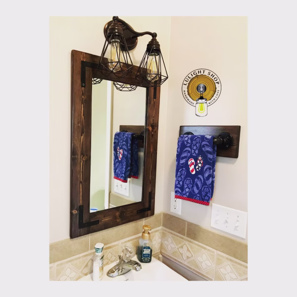 Coffee-stained pine bathroom mirror with dark metal corner brackets above a sink, shown with a cage vanity light and towel holder sold separately.